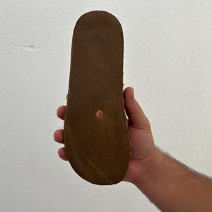 Puede incluir: Suela de sandalia de cuero marr&oacute;n claro, mostrada sobre un fondo blanco. La suela presenta una textura marr&oacute;n oscuro y un peque&ntilde;o remache redondo color cobre en el centro.