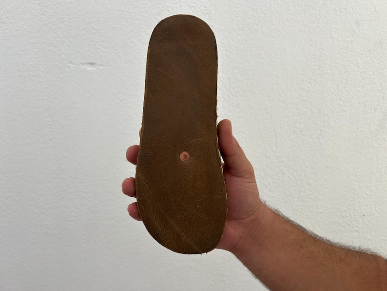 Puede incluir: Suela de sandalia de cuero marr&oacute;n claro, mostrada sobre un fondo blanco. La suela presenta una textura marr&oacute;n oscuro y un peque&ntilde;o remache redondo color cobre en el centro.