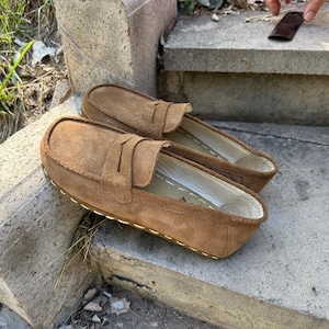 Puede incluir: Un par de mocasines de ante marr&oacute;n con dise&ntilde;o sin cordones. Los zapatos tienen una puntera redondeada y una correa decorativa en la parte superior. Las suelas est&aacute;n cosidas con hilo blanco. Los zapatos est&aacute;n sobre una superficie de hormig&oacute;n.