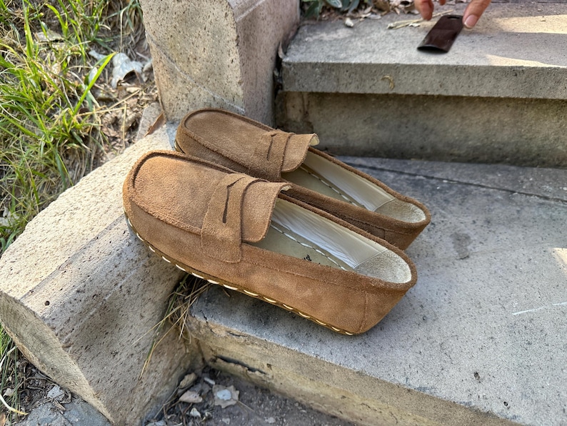 Puede incluir: Un par de mocasines de ante marr&oacute;n con dise&ntilde;o sin cordones. Los zapatos tienen una puntera redondeada y una correa decorativa en la parte superior. Las suelas est&aacute;n cosidas con hilo blanco. Los zapatos est&aacute;n sobre una superficie de hormig&oacute;n.
