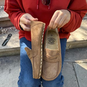 Puede incluir: Un par de mocasines de ante marr&oacute;n con costuras blancas y un dise&ntilde;o de mocas&iacute;n con borlas. Los zapatos son sostenidos por una persona que lleva una sudadera con capucha roja y vaqueros azules. El texto "handmade barefoot" est&aacute; en el interior.