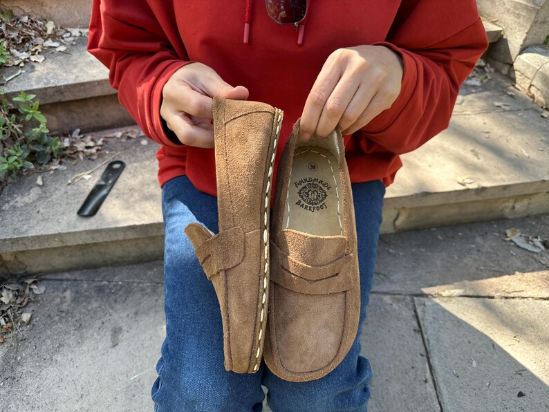 Puede incluir: Un par de mocasines de ante marr&oacute;n con costuras blancas y un dise&ntilde;o de mocas&iacute;n con borlas. Los zapatos son sostenidos por una persona que lleva una sudadera con capucha roja y vaqueros azules. El texto "handmade barefoot" est&aacute; en el interior.