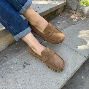 Puede incluir: Mocasines de ante marr&oacute;n claro con dise&ntilde;o cl&aacute;sico de penny loafer. Los zapatos son de color marr&oacute;n claro y tienen una suela cosida. La persona lleva vaqueros azules con los pu&ntilde;os doblados.
