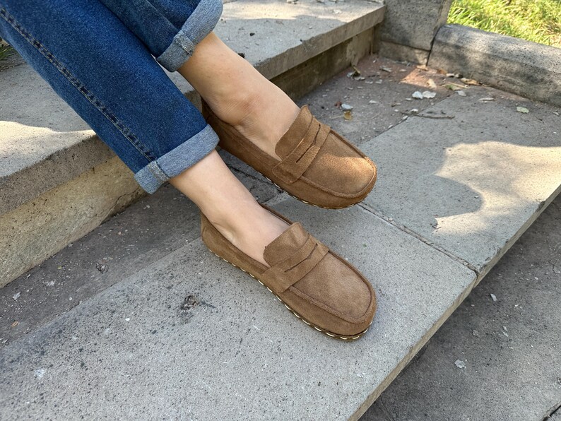 Puede incluir: Mocasines de ante marr&oacute;n claro con dise&ntilde;o cl&aacute;sico de penny loafer. Los zapatos son de color marr&oacute;n claro y tienen una suela cosida. La persona lleva vaqueros azules con los pu&ntilde;os doblados.