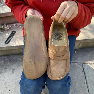 Puede incluir: Mocasines de ante marr&oacute;n con suelas cosidas. El zapato derecho presenta una correa decorativa y el texto "HANDMADE BAREFOOT" en el interior. Los zapatos se sostienen sobre un fondo de vaqueros azules y una sudadera con capucha roja.