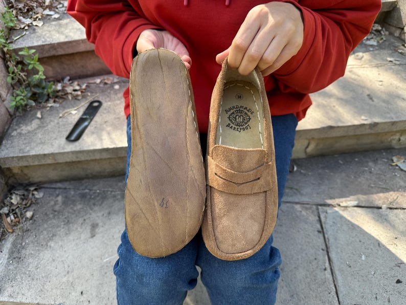 Puede incluir: Mocasines de ante marr&oacute;n con suelas cosidas. El zapato derecho presenta una correa decorativa y el texto "HANDMADE BAREFOOT" en el interior. Los zapatos se sostienen sobre un fondo de vaqueros azules y una sudadera con capucha roja.