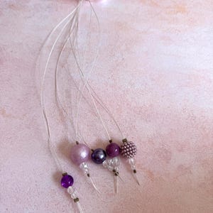 Puede incluir: Una colección de adornos de cuentas en tonos morados, lavanda y transparentes. Cada adorno presenta un diseño de cuentas diferente, ensartado en un fino alambre plateado y atado con una cinta rosa. Las cuentas varían en tamaño y textura.