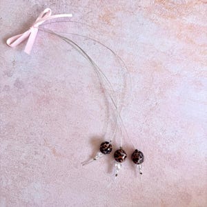 Puede incluir: Tres juguetes para gatos con cuentas con estampado de leopardo, unidos a finos alambres y un lazo de cinta rosa. Las cuentas son marrones y negras, sobre un fondo rosa claro.