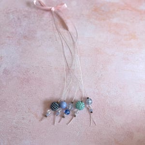 Puede incluir: Una colección de abalorios con cuentas en tonos azules y transparentes, unidos a finos cordones plateados. Cada abalorio presenta un diseño de cuentas diferente, incluyendo esferas texturizadas y lisas. Una pequeña cinta rosa está atada en la parte superior.