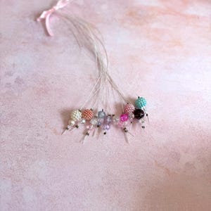 Puede incluir: Un conjunto de horquillas para el pelo con cuentas de varios colores, incluyendo blanco, rosa y turquesa. Cada horquilla presenta un grupo de cuentas y está unida a un alfiler plateado. Una pequeña cinta rosa está atada en la parte superior.