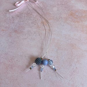 Puede incluir: Un accesorio para el cabello hecho a mano con cuentas y cristales azules. El diseño incluye una cuenta redonda central, una cuenta texturizada y pequeños detalles de cristal. Una delicada cinta rosa está atada en la parte superior.