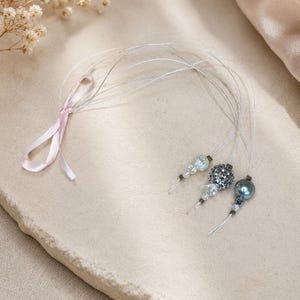 May include: Three beaded hair accessories with clear, silver, and dark blue beads. Each accessory has a small cluster of beads attached to a thin, clear wire. A pink ribbon is tied to the wires. The accessories are on a light beige surface.