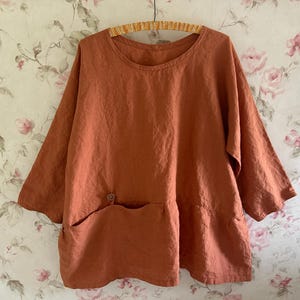 May include: A loose-fitting, rust-colored top with a round neckline and three-quarter length sleeves. The garment features a large, horizontal front pocket positioned across the lower section, accented with a single small button detail.