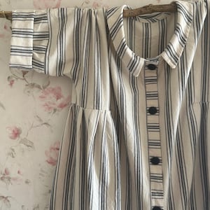 May include: A cream-coloured dress with vertical navy blue stripes. It has a rounded collar, short sleeves, and a button-down front with black buttons. The dress is hanging on a wooden rod against a floral wallpaper background.