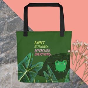 May include: Green tote bag with a frog illustration and the text "Expect nothing. Appreciate everything." printed on the front. The bag has black handles and a green and white leafy design.