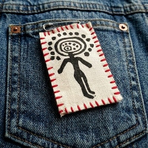 Könnte beinhalten: Ein rechteckiger Aufnäher mit einem schwarzen Figuren-Design, roten Nähten und einer Sicherheitsnadel, die an einer Jeans-Tasche befestigt ist. Die Figur hat einen kreisförmigen Kopf und ausgestreckte Arme und Beine. Der Aufnäher ist aus hellem Stoff.