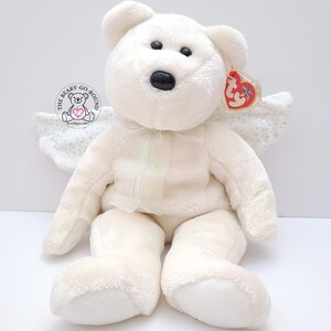 Op de afbeelding: Een witte teddybeer met engelen vleugels en een rood hartvormig label. De beer heeft zwarte ogen en een zwarte neus. Hij heeft een cr&egrave;mekleurige strik en een cr&egrave;mekleurig paneel op de borst. De vleugels zijn wit met zilveren glitter.