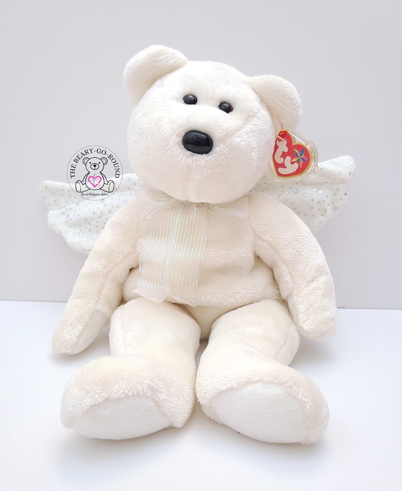 Op de afbeelding: Een witte teddybeer met engelen vleugels en een rood hartvormig label. De beer heeft zwarte ogen en een zwarte neus. Hij heeft een cr&egrave;mekleurige strik en een cr&egrave;mekleurig paneel op de borst. De vleugels zijn wit met zilveren glitter.