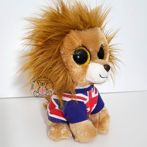 May include: A plush lion toy with large, glittery eyes and a shaggy mane. The lion is wearing a blue and red shirt with a Union Jack design. The Beary Go Round logo is visible on the left side.