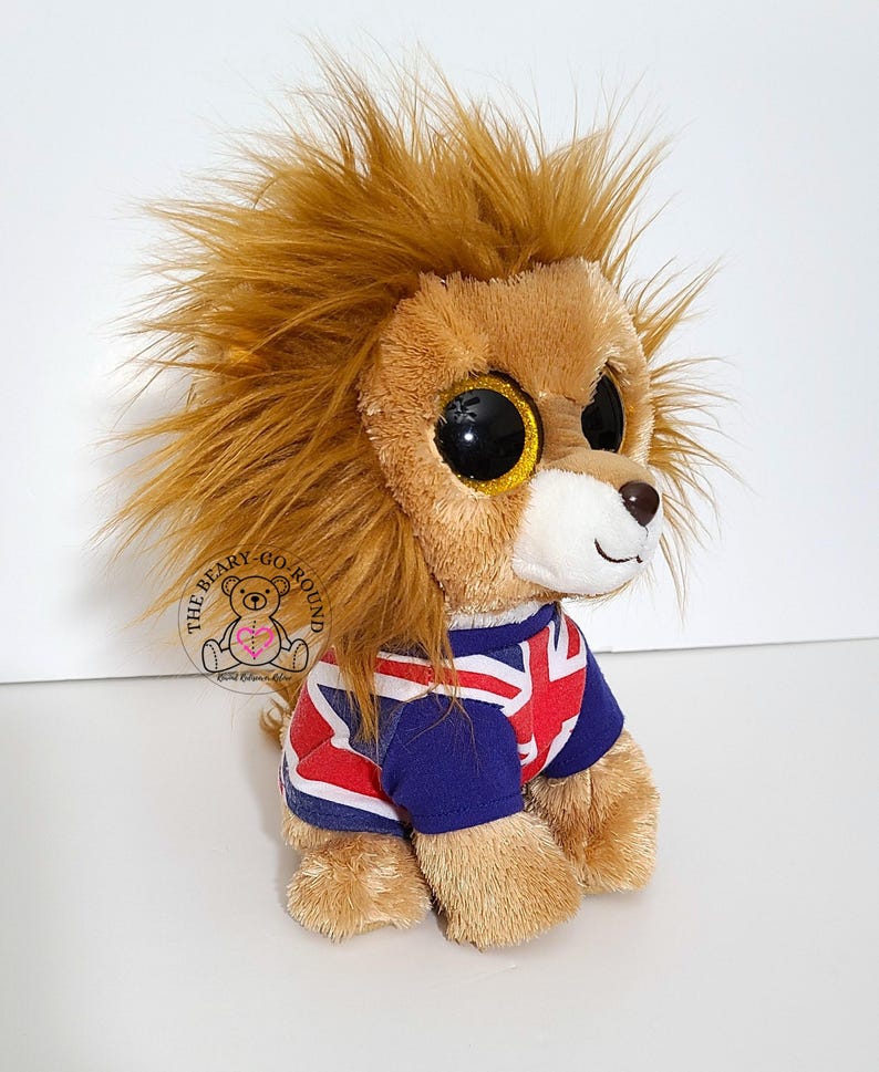 May include: A plush lion toy with large, glittery eyes and a shaggy mane. The lion is wearing a blue and red shirt with a Union Jack design. The Beary Go Round logo is visible on the left side.