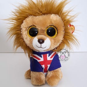 May include: A plush lion toy with large, glittery eyes and a brown mane. The lion wears a blue shirt with a Union Jack flag design. A heart-shaped tag is attached to the toy.
