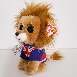 May include: A plush lion toy with large, glittery eyes and a brown mane. The lion wears a blue and white shirt with a Union Jack design. A red heart-shaped tag is attached to its ear.