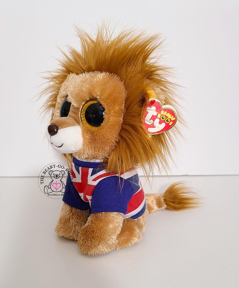 May include: A plush lion toy with large, glittery eyes and a brown mane. The lion wears a blue and white shirt with a Union Jack design. A red heart-shaped tag is attached to its ear.