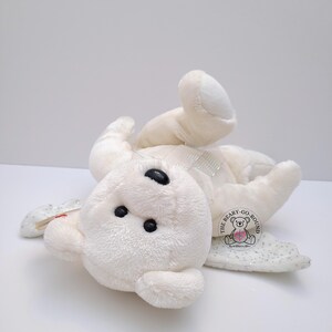 Op de afbeelding: Een witte teddybeer met zwarte ogen en een doorzichtige strik. De beer ligt op zijn rug met zijn armen en benen uitgestrekt. Een label met de tekst "THE BEARY-GO-ROUND" is bevestigd.