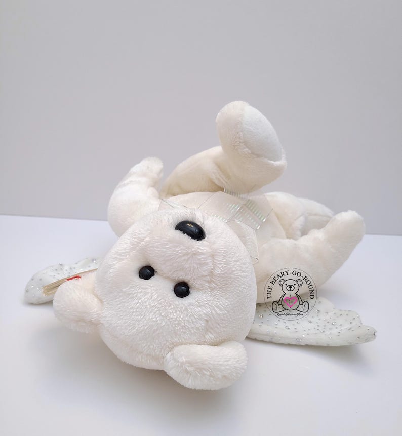 Op de afbeelding: Een witte teddybeer met zwarte ogen en een doorzichtige strik. De beer ligt op zijn rug met zijn armen en benen uitgestrekt. Een label met de tekst "THE BEARY-GO-ROUND" is bevestigd.