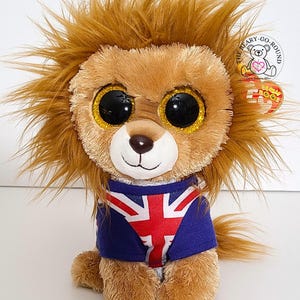May include: A plush lion toy with a brown mane, large black eyes with gold glitter accents, and a blue shirt featuring the Union Jack flag. The toy has a white face and a small brown nose. A tag is attached to the toy.