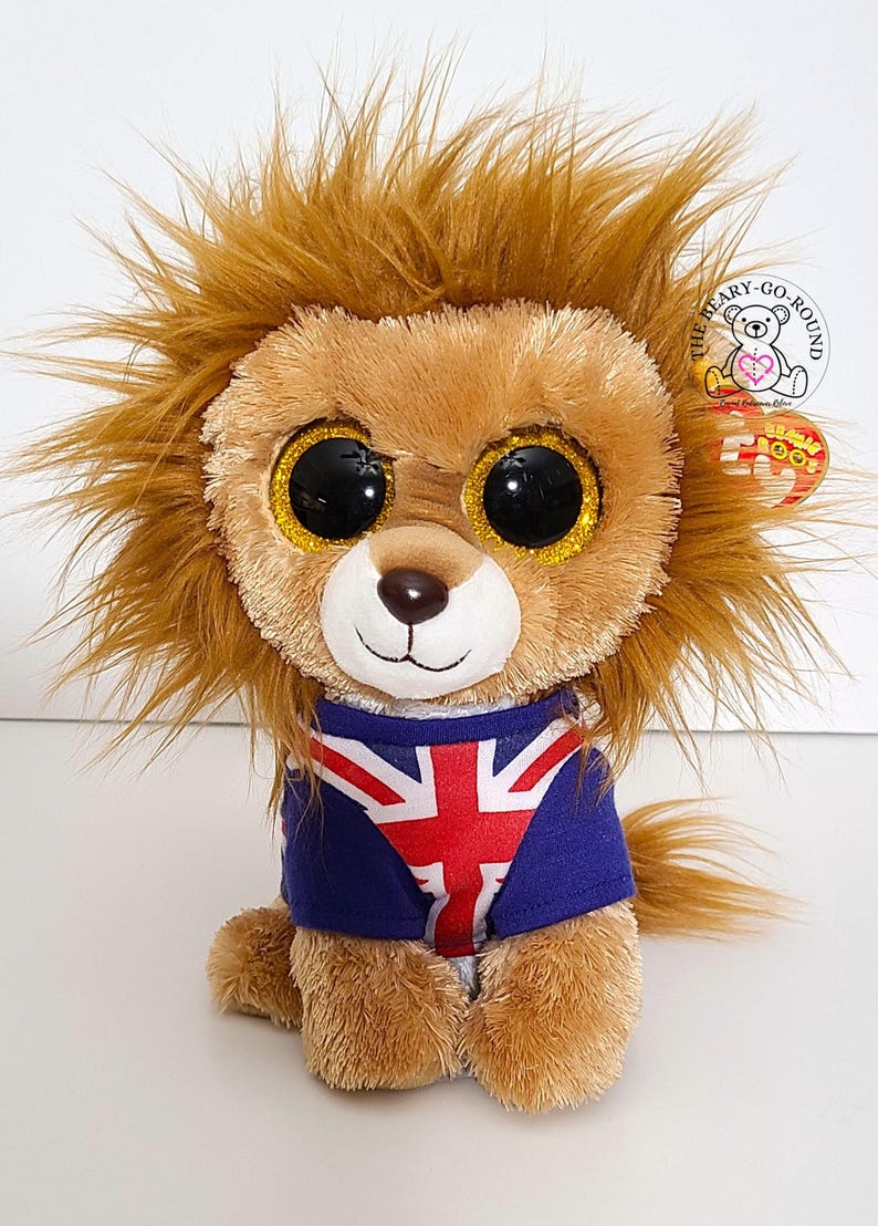 May include: A plush lion toy with a brown mane, large black eyes with gold glitter accents, and a blue shirt featuring the Union Jack flag. The toy has a white face and a small brown nose. A tag is attached to the toy.