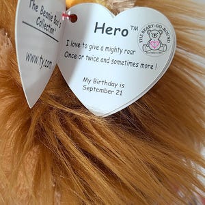 May include: A plush lion toy from The Beanie Boo's Collection, featuring a mane of golden-brown fur. The toy has a heart-shaped tag that reads "Hero" and includes the text "I love to give a mighty roar." The lion's birthday is September 21.