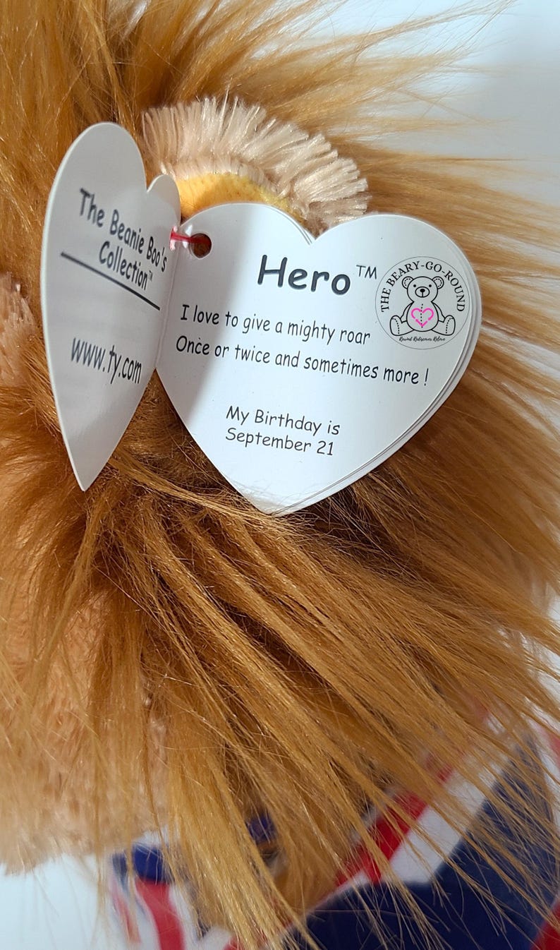 May include: A plush lion toy from The Beanie Boo's Collection, featuring a mane of golden-brown fur. The toy has a heart-shaped tag that reads "Hero" and includes the text "I love to give a mighty roar." The lion's birthday is September 21.