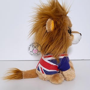 May include: A plush lion toy with a brown mane and tail, wearing a blue and red Union Jack t-shirt. The lion has a white muzzle and a small, round, black eye. The toy is sitting on a white surface.
