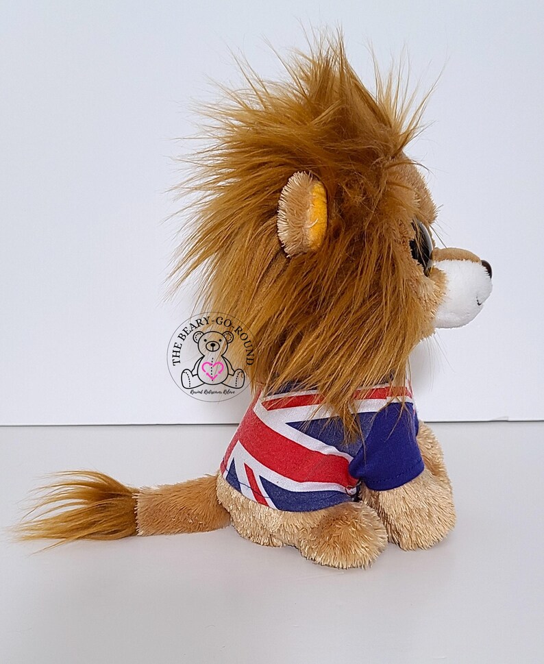 May include: A plush lion toy with a brown mane and tail, wearing a blue and red Union Jack t-shirt. The lion has a white muzzle and a small, round, black eye. The toy is sitting on a white surface.
