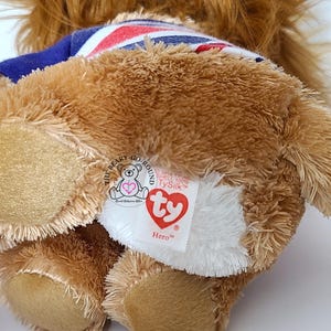 May include: A brown plush bear toy with a blue and Union Jack patterned collar. The bear has a white patch with a red heart-shaped tag that reads "Hero". The bear is a Ty Beanie Baby.