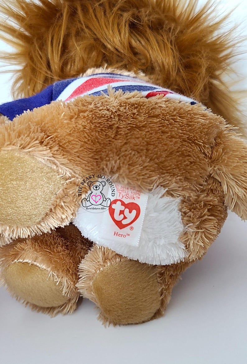 May include: A brown plush bear toy with a blue and Union Jack patterned collar. The bear has a white patch with a red heart-shaped tag that reads "Hero". The bear is a Ty Beanie Baby.