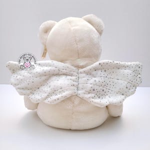 Op de afbeelding: Een cr&egrave;mekleurige pluche teddybeer met witte engelen vleugels. De vleugels zijn versierd met zilveren pailletten. De beer heeft een label met de tekst "The Beary-Go-Round". De beer staat op een wit oppervlak.