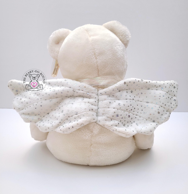 Op de afbeelding: Een cr&egrave;mekleurige pluche teddybeer met witte engelen vleugels. De vleugels zijn versierd met zilveren pailletten. De beer heeft een label met de tekst "The Beary-Go-Round". De beer staat op een wit oppervlak.