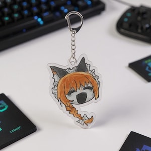 Puede incluir: Un llavero acr&iacute;lico transparente con un personaje de dibujos animados con cabello naranja y una trenza. El personaje tiene un tocado blanco y negro y est&aacute; unido a una cadena y un cierre plateados. Un accesorio de juego.