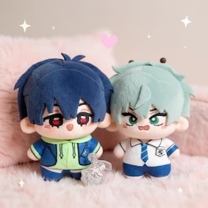 May include: Two plush dolls with anime-style features. One doll has blue hair and a blue and green outfit, the other has mint green hair and a white and blue uniform. Both dolls have detailed facial features and are standing on a pink surface.