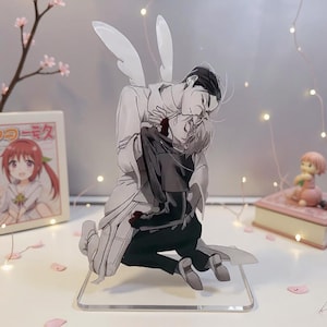 May include: A clear acrylic stand featuring a manga-style illustration of two figures embracing. One figure, with white wings, wears a white outfit, while the other is in black. The artwork is set against a soft background with fairy lights and a small framed image.