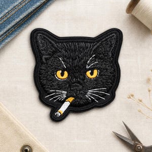 May include: An embroidered patch featuring the head of a black cat with bright yellow eyes and white whiskers. The cat is depicted with a cigarette in its mouth, rendered in white and orange thread, with a black stitched border outline.