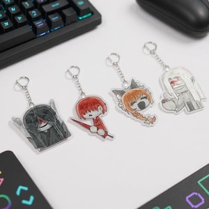 Puede incluir: Cuatro llaveros de estilo anime con cadenas y anillos plateados. Cada llavero presenta un dise&ntilde;o de personaje diferente, incluido un personaje con cabello rojo y una espada, y un personaje con cabello negro largo.