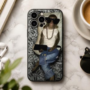 May include: A rectangular smartphone case featuring a graphic print of a monkey wearing black sunglasses, a white tank top, a gold chain, and blue jeans. The background of the print consists of a repeating pattern of United States hundred-dollar bills.