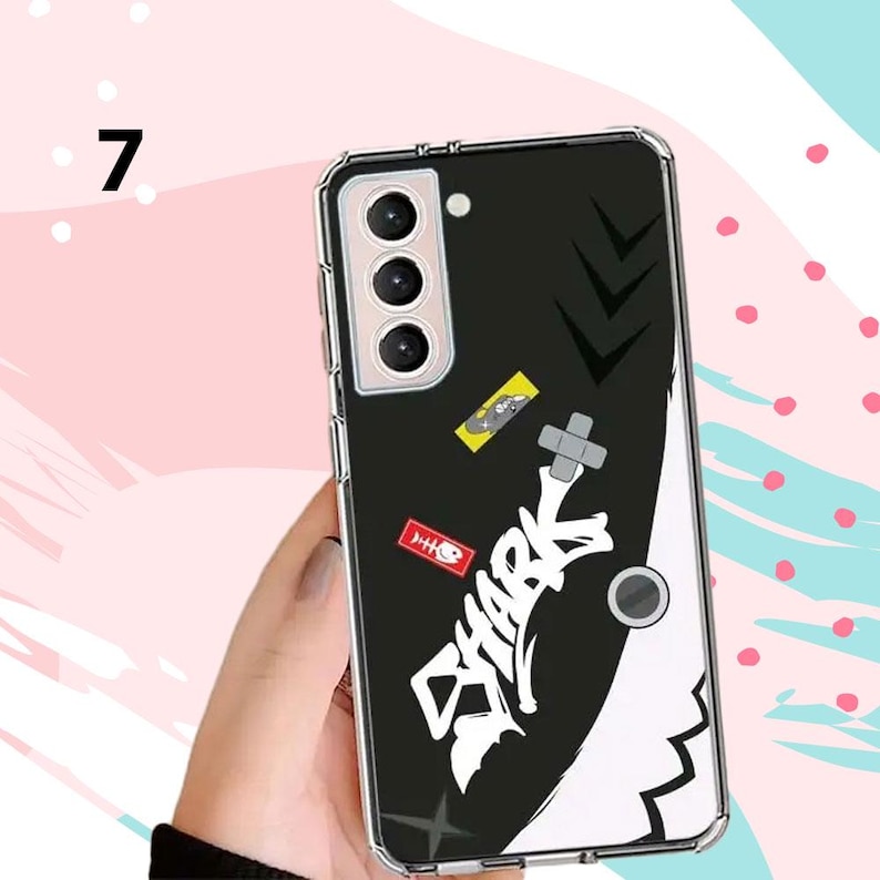 Puede incluir: Funda de tel&eacute;fono negra con un gr&aacute;fico de tibur&oacute;n blanco y la palabra "SHARK" en estilo graffiti. La funda tiene un borde transparente y presenta peque&ntilde;as pegatinas con varios dise&ntilde;os. La funda del tel&eacute;fono se sostiene en una mano.