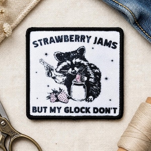 May include: A square embroidered patch with a white background and black border. It features a black and white illustration of a raccoon holding a handgun and a jar of jam, with two strawberries. Text reads: STRAWBERRY JAMS BUT MY GLOCK DON'T.