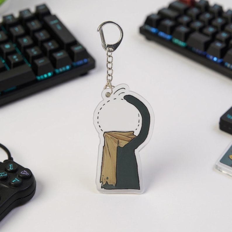 Puede incluir: Un llavero acr&iacute;lico transparente con un gato negro que lleva una bufanda beige. El gato est&aacute; estirando la pata hacia un c&iacute;rculo punteado. El llavero tiene una cadena y un cierre plateados. Teclados y un mando de juegos en el fondo.
