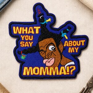 Könnte beinhalten: Ein bestickter Aufnäher mit blauem Hintergrund, der eine Zeichentrickfigur mit schwarzen Zöpfen, blauen Schnullern und dem gelben Text WHAT YOU SAY ABOUT MY MOMMA!? zeigt. Die Figur hat braune Haut und einen weit aufgerissenen Blick.