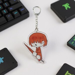 Puede incluir: Un llavero de acr&iacute;lico transparente con un personaje de dibujos animados con pelo rojo y un atuendo rojo sosteniendo un paraguas rojo. El llavero tiene una cadena y un cierre plateados. El fondo incluye un teclado negro y objetos cuadrados negros.